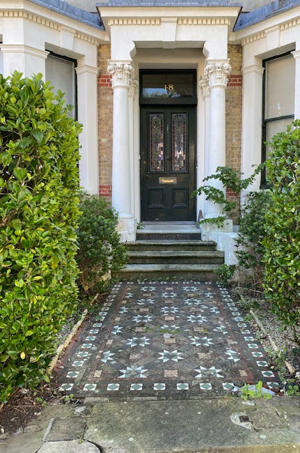 A serene garden scene featuring a neatly trimmed green lawn bordered by a variety of lush plants and shrubs. To the left, there is a light-colored wooden garden bench placed on a small paved area, sur